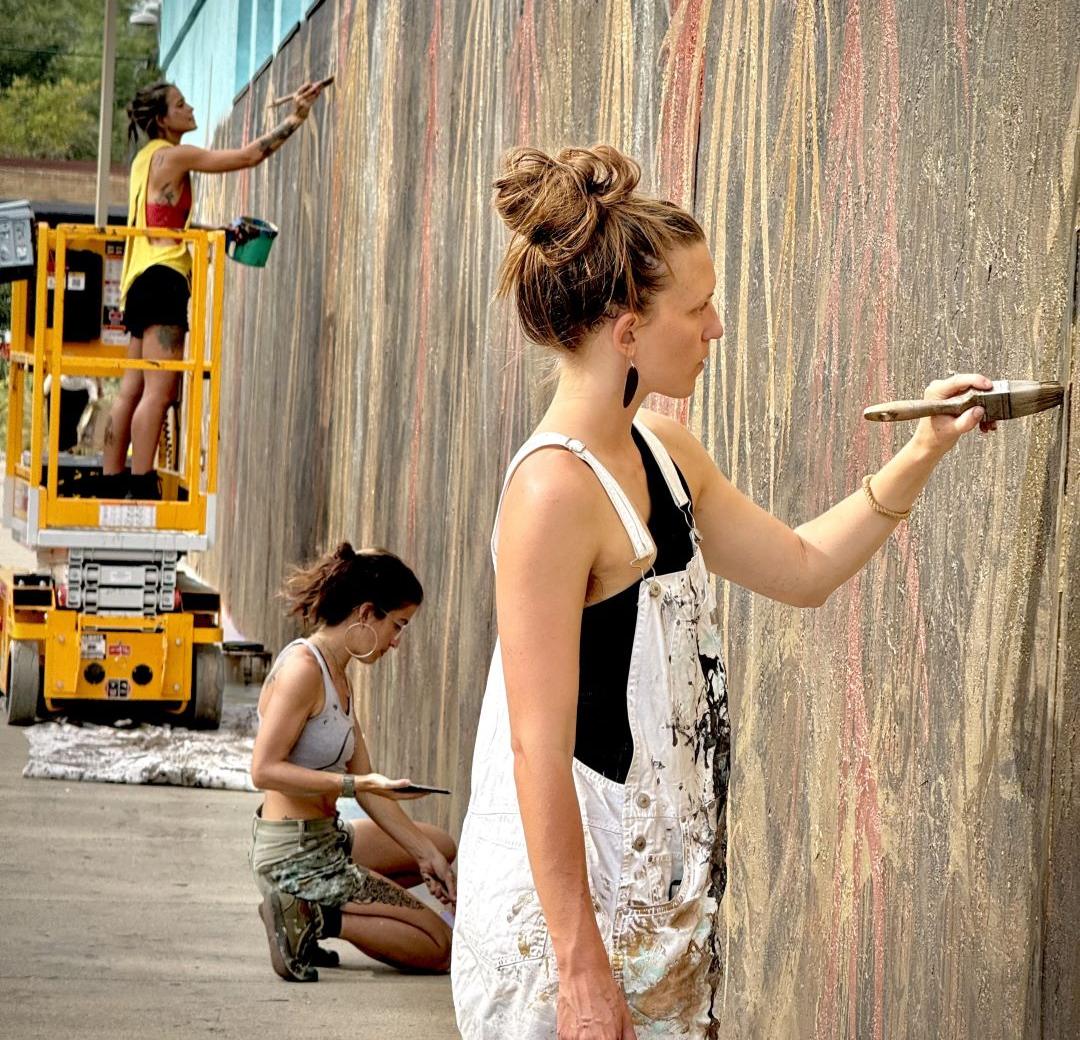 Three women in painting clothes paint the mural at the Willy Street Co-Op