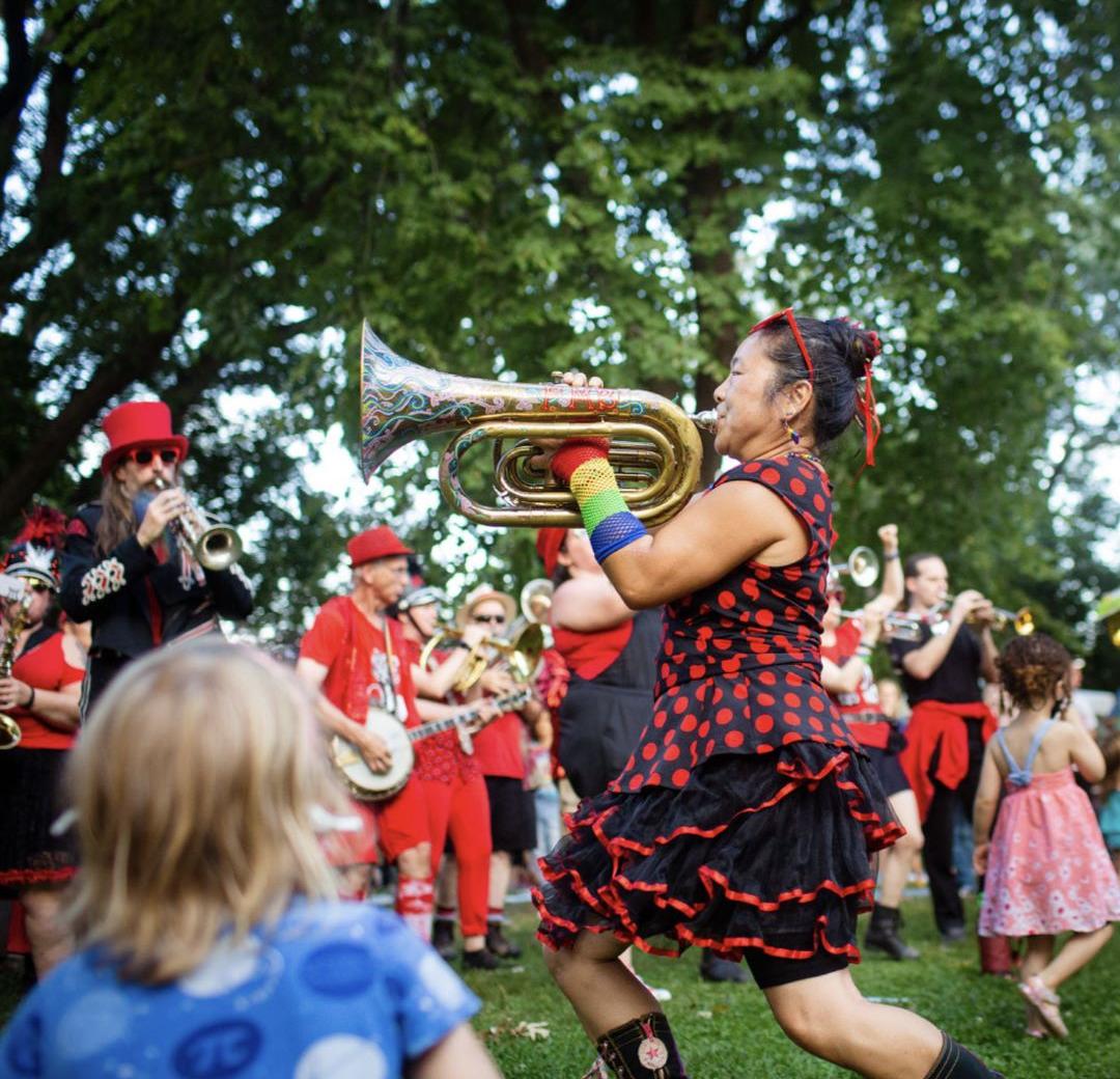 A colorfully dressed woman plays a large trumpet surrounded by other musicians and spectators